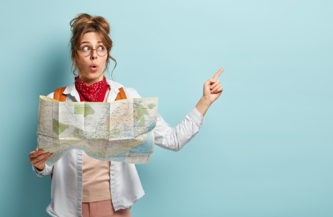 Shocked impressed female tourist points at copy space, holds travel map, searches right route during trip, surprised to be lost in unknown place, isolated on blue wall. Traveling and tourism concept