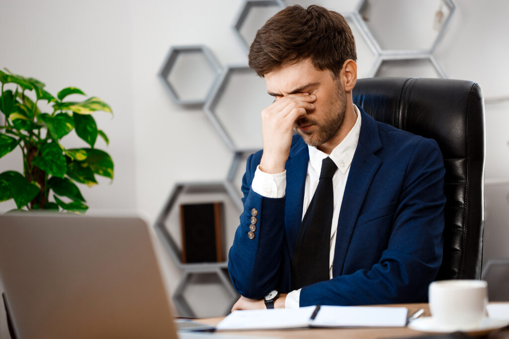 Upset young businessman sitting at workplace, office background.